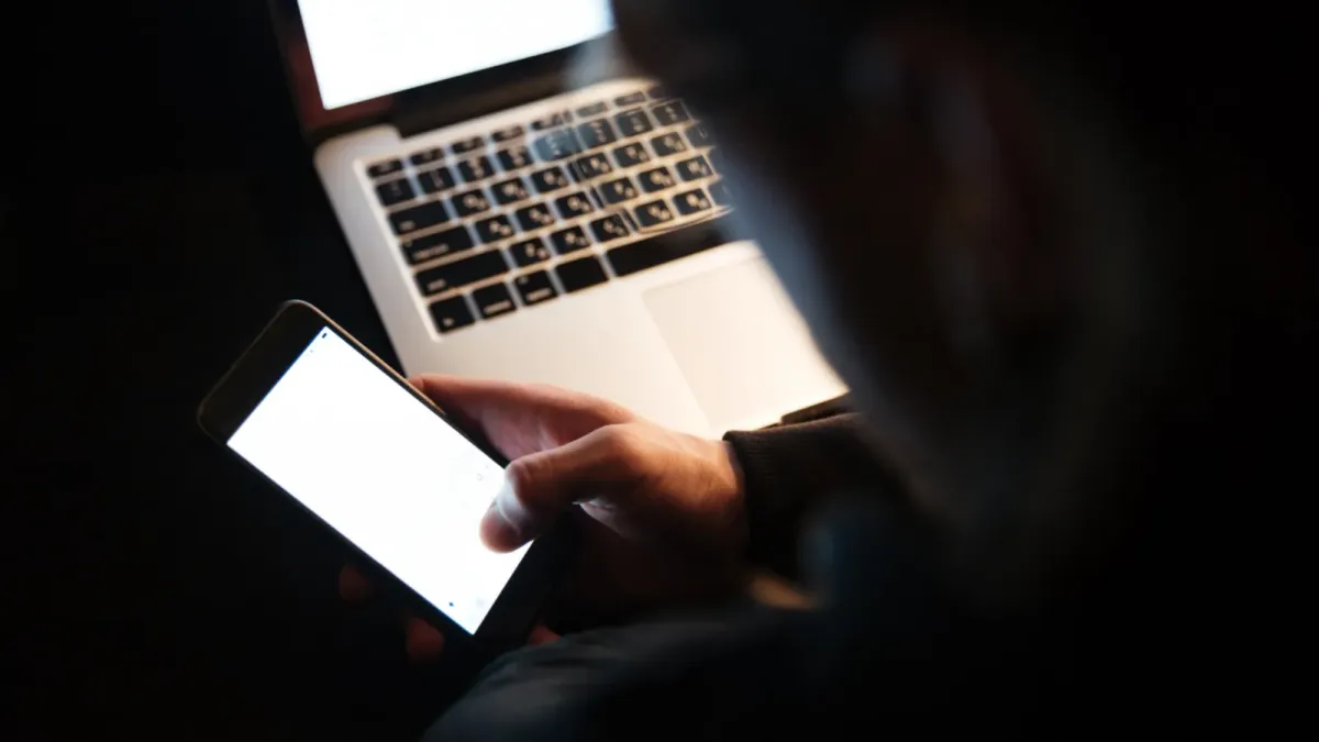 Muž v přítmí drží chytrý telefon nad klávesnicí notebooku