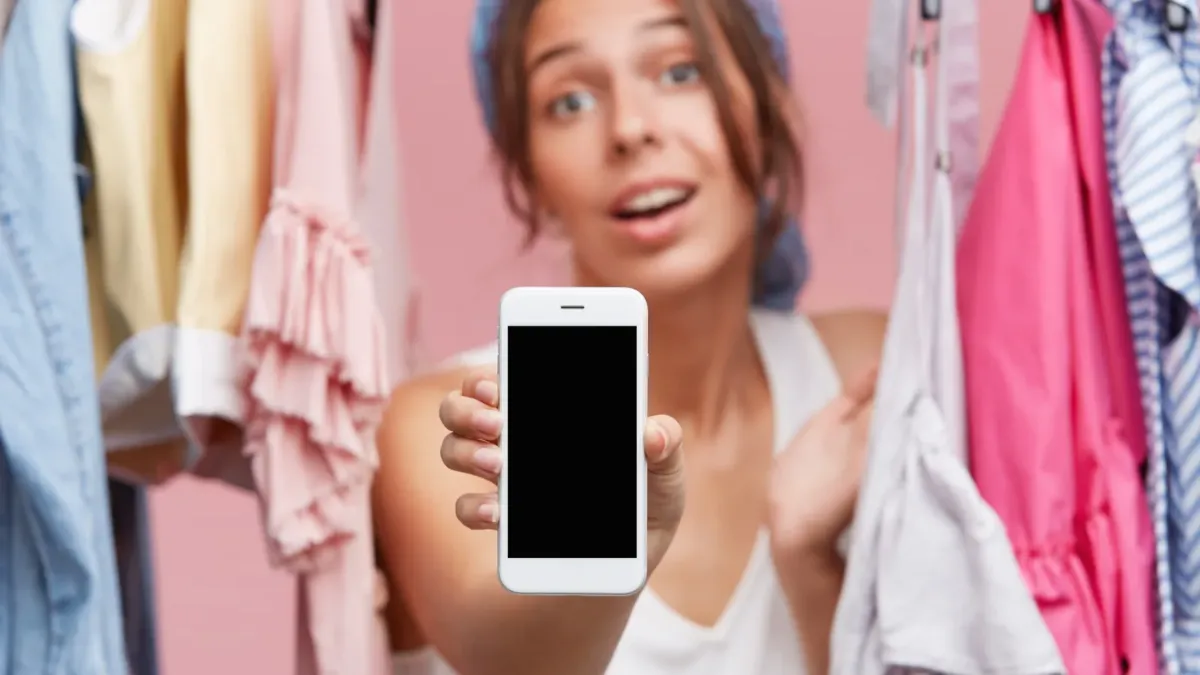 Žena ukazuje mobilní telefon před věšákem s oblečením na prodej