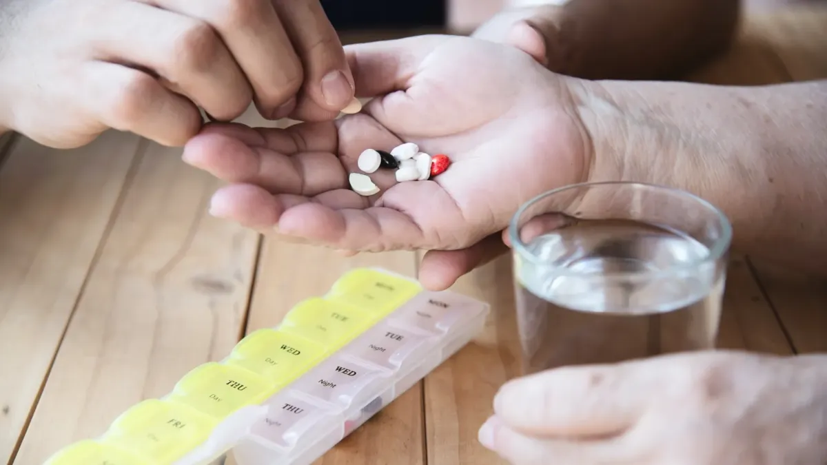 Starší člověk bere tablety z týdenního dávkovače vedle sklenice vody